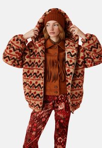 Flauschige Jacke in Braun- und Crememuster mit Kapuze, getragen über einem gefransten hellbraunen Hemd, kombiniert mit rotblumigen Hose und einer passenden Strickmütze.