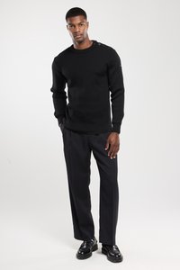 Hombre vestido con un suéter negro con botones en los hombros, pantalones negros y zapatos negros, de pie con las manos parcialmente en los bolsillos sobre un fondo blanco.