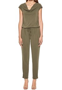 Kobieta w oliwkowym kombinezonie z krótkim rękawem, z dekoltem drapowanym, z talią na sznurek i beżowych sandałach na obcasie, stojąca na białym tle.