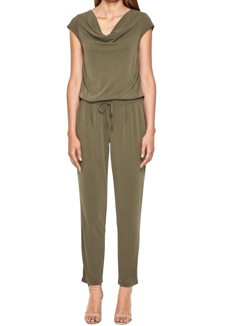 Kobieta w oliwkowym kombinezonie z krótkim rękawem, z dekoltem drapowanym, z talią na sznurek i beżowych sandałach na obcasie, stojąca na białym tle.