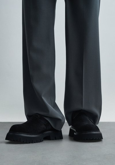 Pantofi platform din suede negru, cu talpă groasă, purtați cu pantaloni gri largi, pe un podea gri, împotriva unui fundal simplu.