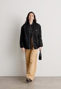 Sort bomberjakke med lynlås foran og brystlomme, parret med beige brede bukser. Modellen bærer en lille sort håndtaske og har sorte sko på.