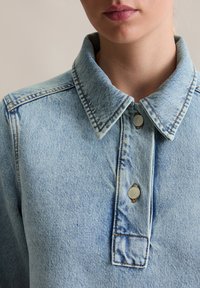 Haut en denim bleu clair avec un col pointu, deux boutons-pression argentés et des détails de couture visibles sur le tissu.