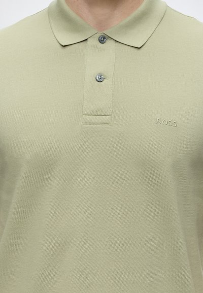 Polo vert clair avec deux boutons et petit logo BOSS brodé sur la poitrine gauche porté par une personne.