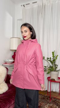 Γυναίκα με ροζ μπουφάν με κουκούλα και μαύρο παντελόνι, στέκεται μέσα σε εσωτερικό χώρο κοντά σε κόκκινο πλαϊνό τραπεζάκι με γλάστρα και άσπρες κουρτίνες στο φόντο.