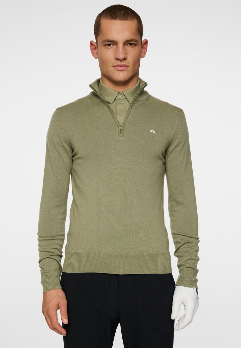 Olivgrön stickad tröja med dragkedja i kragen, långa ärmar, ribbade manschetter och nederkant, med en subtil logotyp på bröstet.