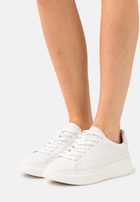 Baskets en cuir blanc avec des lacets plats, bout rond et semelle légèrement surélevée. Texture lisse et design minimaliste.