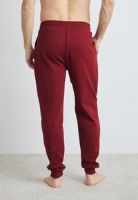 Rote Jogginghose mit schmal zulaufendem Bein, elastischem Bund und seitlichen Taschen, aus weichem Stoff, von hinten auf einem neutralen Hintergrund gezeigt.