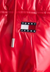 Červená lesklá bunda s viazaním, so štvorcovou čiernou nášivkou s nápisom "TOMMY JEANS" a trojfarebnou etiketou v červenej, bielej a modrej farbe.