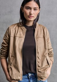 Giubbotto bomber in suede marrone chiaro con chiusura a zip, due tasche laterali e polsini a costine, indossato sopra una camicia marrone scuro. Jeans in denim completano il look.