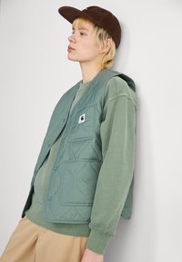 Gilet verde trapuntato con una texture liscia, dotato di tasca a toppa e chiusure a pressione, indossato sopra un pullover verde abbinato e pantaloni beige.