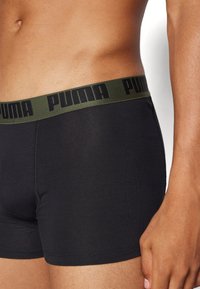Boxer shorts pretos com uma cintura elástica verde, apresentando "PUMA" em letras pretas. O tecido parece suave com um design ajustado.