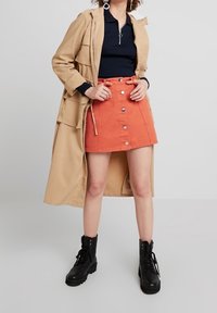 Abrigo largo beige sobre un polo azul marino, combinado con una falda de mezclilla naranja brillante con botones. Botas de combate negras completan el atuendo.