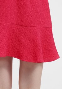 Ourlet de robe rouge texturé avec motif pied-de-poule, coupe évasée, portée au-dessus de jambes nues sur un fond clair uni.