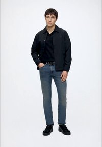 Junger Mann steht mit einer Hand in der Tasche, trägt schwarze Jacke, schwarzes Hemd, blaue Jeans, schwarzen Gürtel und schwarze Schuhe vor weißem Hintergrund.