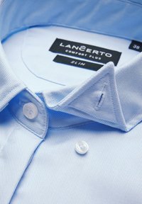 Jasnoniebieska, dopasowana koszula z teksturowanej tkaniny. Posiada kołnierz w kształcie ostrego oraz dwa guziki, a także metkę z napisem "LANCERTO Comfort Plus."