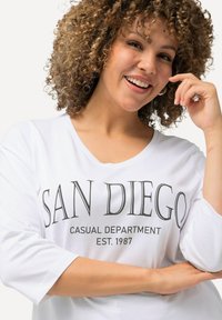 Weißes, locker sitzendes T-Shirt mit V-Ausschnitt und 3/4-Ärmeln. Auf der Vorderseite befindet sich der schwarze Text "SAN DIEGO CASUAL DEPARTMENT EST. 1987".