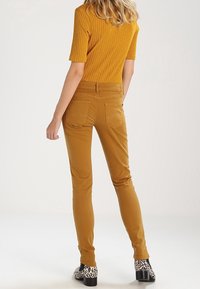 Slim-fit mustard yellow pants with a smooth texture, back pockets, and a mid-rise waist, paired with black and white patterned heels.