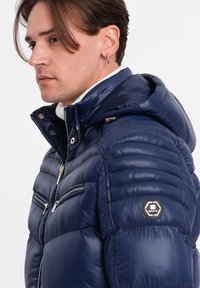 Granatowa kurtka puchowa z kapturem na zamek, pikowana konstrukcja oraz dwie kieszenie z przodu na zamek. Posiada naszywkę z logo na lewym rękawie.