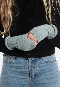 Guanti senza dita a maglia azzurro chiaro con texture a coste, indossati su una giacca in corduroy nera, che mostrano un design aderente e un accento in pelle.