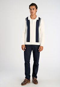 Νεαρός άνδρας που στέκεται, φοράει λευκό πλεκτό πουλόβερ με ναυτικά κάθετα ρίγες, σκούρο παντελόνι και καφέ δερμάτινα παπούτσια με κορδόνια, μπροστά σε απλό φόντο.