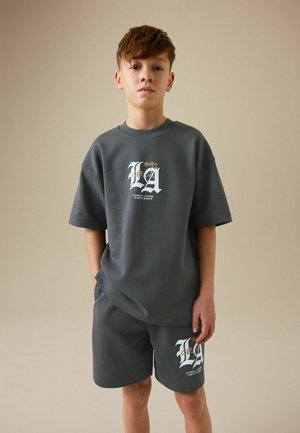 Dreng står med hænderne i lommerne iført mørkegrå oversized T-shirt og matchende shorts med hvidt "LA" tekst- og koordinatdesign.
