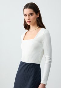 Top a maniche lunghe rigato bianco con scollatura quadrata, abbinato a una gonna scura a righe sottili. L'outfit è completato da una collana di perle e orecchini pendenti.