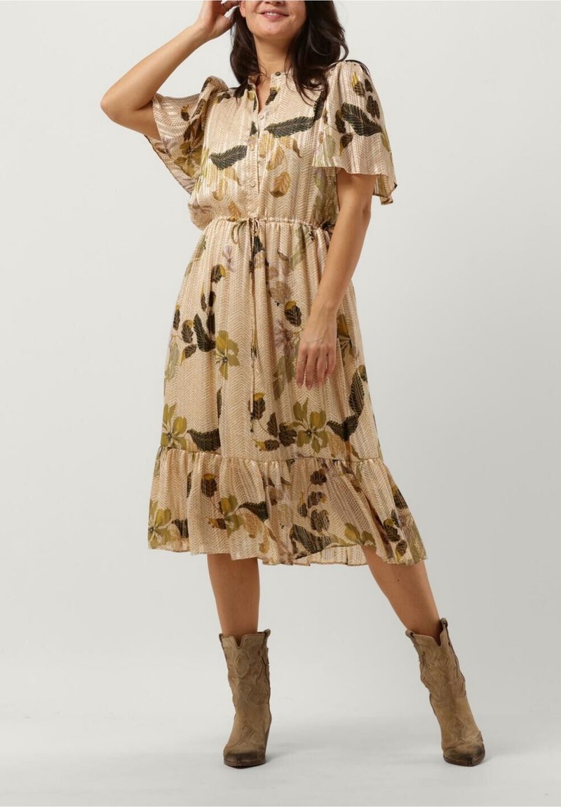 Femme portant une robe midi beige à motifs floraux avec des manches volantes et des bottes de cow-boy marron, posant avec une main près du visage.
