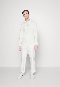 White hoodie with drawstring, front pocket, and logo. Paired with rolled-up white pants and white sneakers, standing on a neutral background.