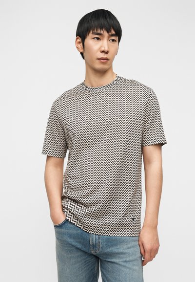 Κοντομάνικο t-shirt σε μπεζ χρώμα με μαύρο ζιγκ ζαγκ μοτίβο, στρογγυλή λαιμόκοψη και άνετη γραμμή, συνδυασμένο με μπλε τζιν.