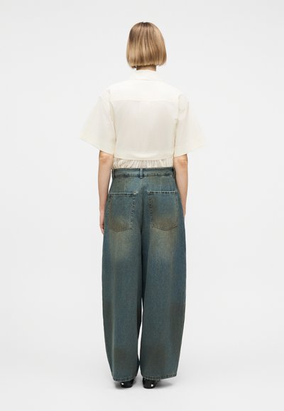 Άτομο με κοντά ξανθά μαλλιά που φοράει κρεμ μπλούζα με κοντά μανίκια και φαρδιά ξεθωριασμένα μπλε τζιν παντελόνια, στέκεται πλάτη σε λευκό φόντο.