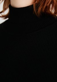 Juodas ripsuotas golfas, kuriame yra aukštas kaklas, tekstūruotas paviršius ir prigludęs dizainas, akcentuojantys vertikalius raštus ir tampumą.