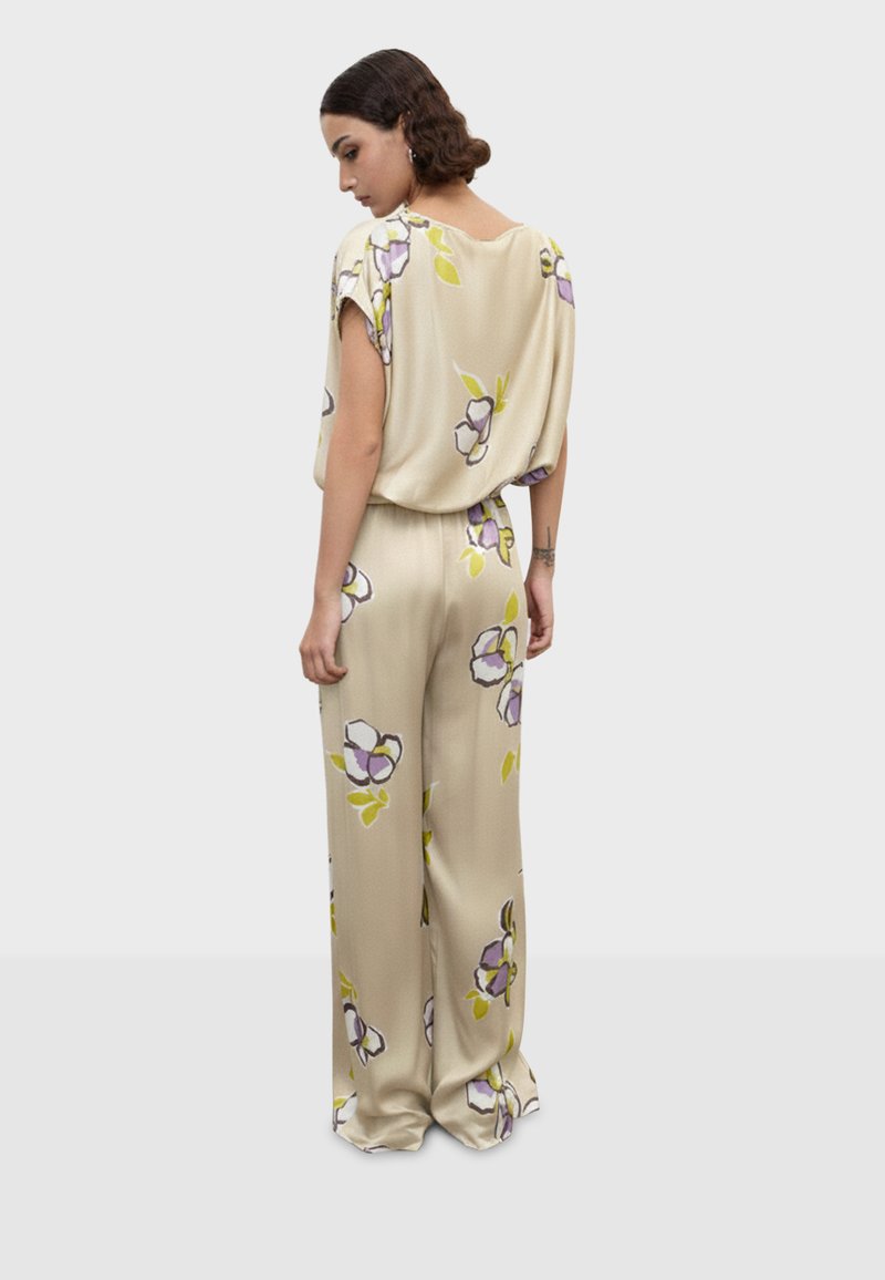 Femme aux cheveux courts et bouclés portant une combinaison ample beige à motifs floraux, debout la tête tournée et les bras détendus le long du corps.