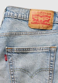 Blugi din denim de un albastru deschis, cu eticheta roșie și patch din piele cu detalii ale brandului, buzunare cusute și fir galben în contrast.