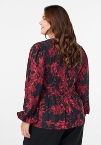 Kvinde med langt brunt hår iført en sort bluse med rød blomsterprint og smockdetaljer, som står sidelæns mod en hvid baggrund.