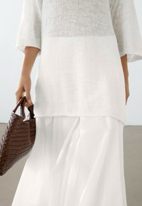 Witte gebreide top met een losse pasvorm en driekwart mouwen, gecombineerd met een vloeiende witte rok. Een donkerbruin geweven handtas wordt in één hand vastgehouden.