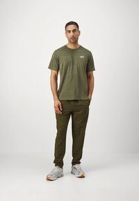 Olivengroen T-shirt met een subtiel patroon en wit logo, gecombineerd met bijpassende taps toelopende broek en grijze sportschoenen met oranje accenten.