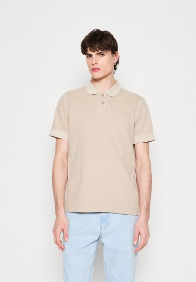 Polo beige en tissu doux, avec un col, un plastron à trois boutons et des manches retroussées. Logo discret imprimé sur la poitrine gauche.