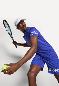 Μπλε αθλητικό μπλουζάκι και σορτσάκι με γράμματα "BORG", κρατώντας μια μπάλα τένις και ρακέτα, φορώντας άσπρο καπέλο. Λείο ύφασμα, αθλητική εφαρμογή.