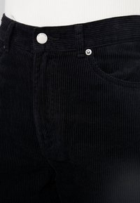 Pantalon en velours côtelé noir avec texture à côtes verticales, design cinq poches et quincaillerie en métal. Gros plan sur la taille et la zone de poche.