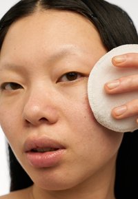 Une personne applique un disque de coton blanc et circulaire sur sa joue, mettant en avant une peau lisse avec une légère texture, une légère rougeur et une couleur de lèvres naturelle.