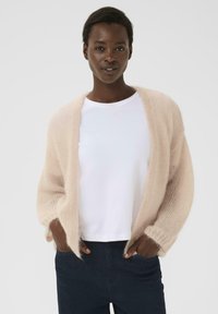 Lys rosa, fluffy cardigan lavet af mohairblanding, åbent frontdesign, lange ærmer, båret over en hvid t-shirt, parret med mørke jeans.