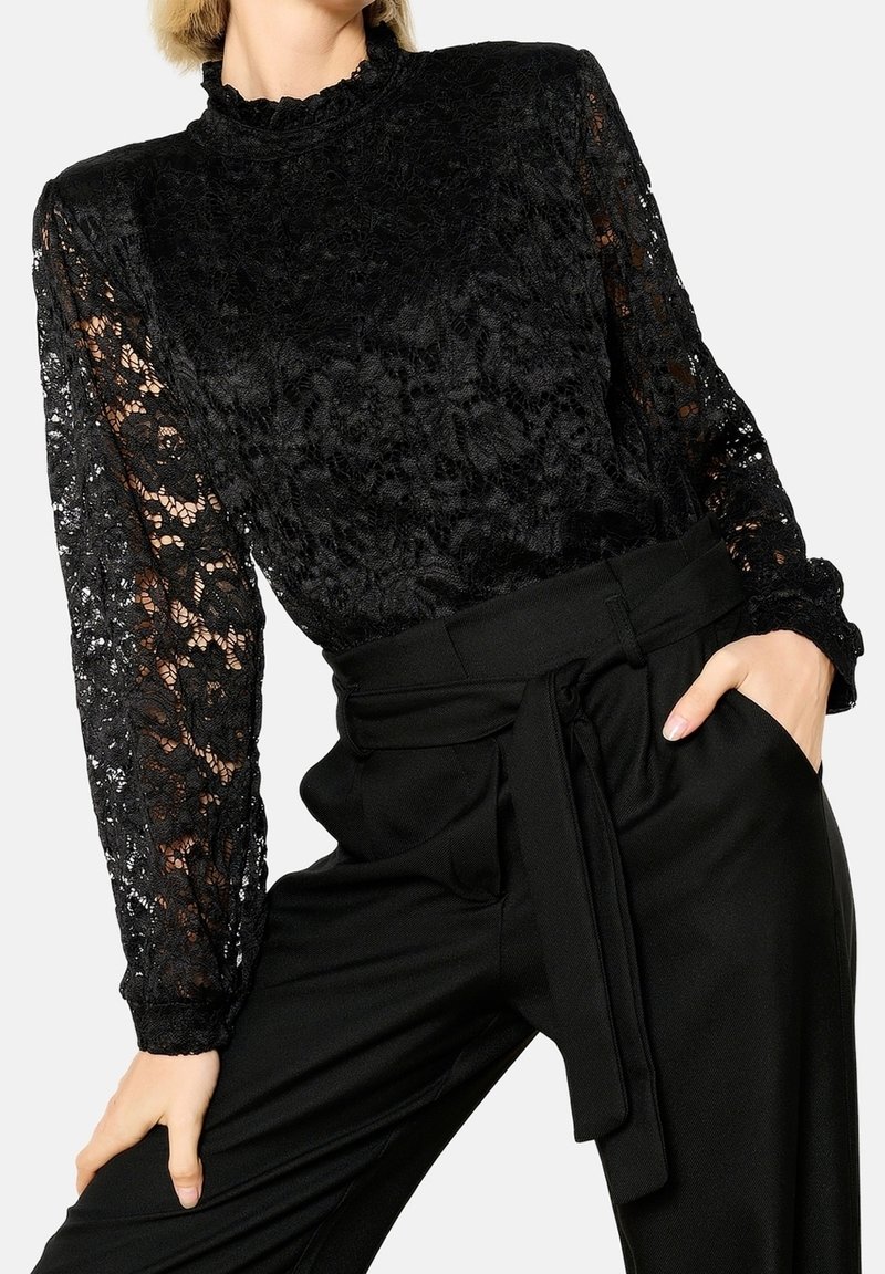 Femme portant un top en dentelle noire à manches longues avec col montant, associé à un pantalon noir taille haute avec une ceinture en tissu nouée à la taille.