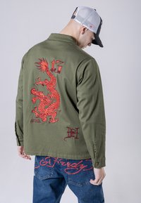 Giacca verde oliva con un grande ricamo di un drago rosso sul retro e un piccolo dettaglio del logo. Il modello indossa jeans blu con accenti rossi.