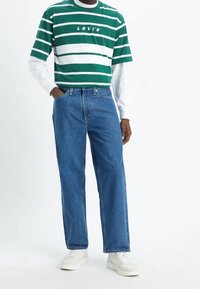 Persona vistiendo una camiseta de rayas verdes y blancas de Levi's sobre una camisa blanca de mangas largas, con jeans azules y zapatillas blancas, de pie con una mano en el bolsillo.