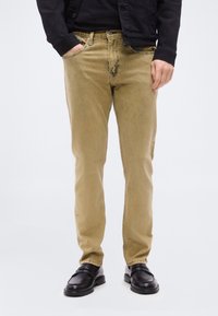 Άνδρας που φοράει μπεζ τζιν στε νής γραμμής, μαύρα loafers, μαύρο πουκάμισο και μαύρο σακάκι με τα χέρια μερικώς στις τσέπες, μπροστά σε απλό λευκό φόντο.