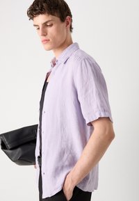 Világos lila, rövid ujjú, lenből készült ing, gombos gallérral és laza szabással, fekete clutch táskával párosítva.