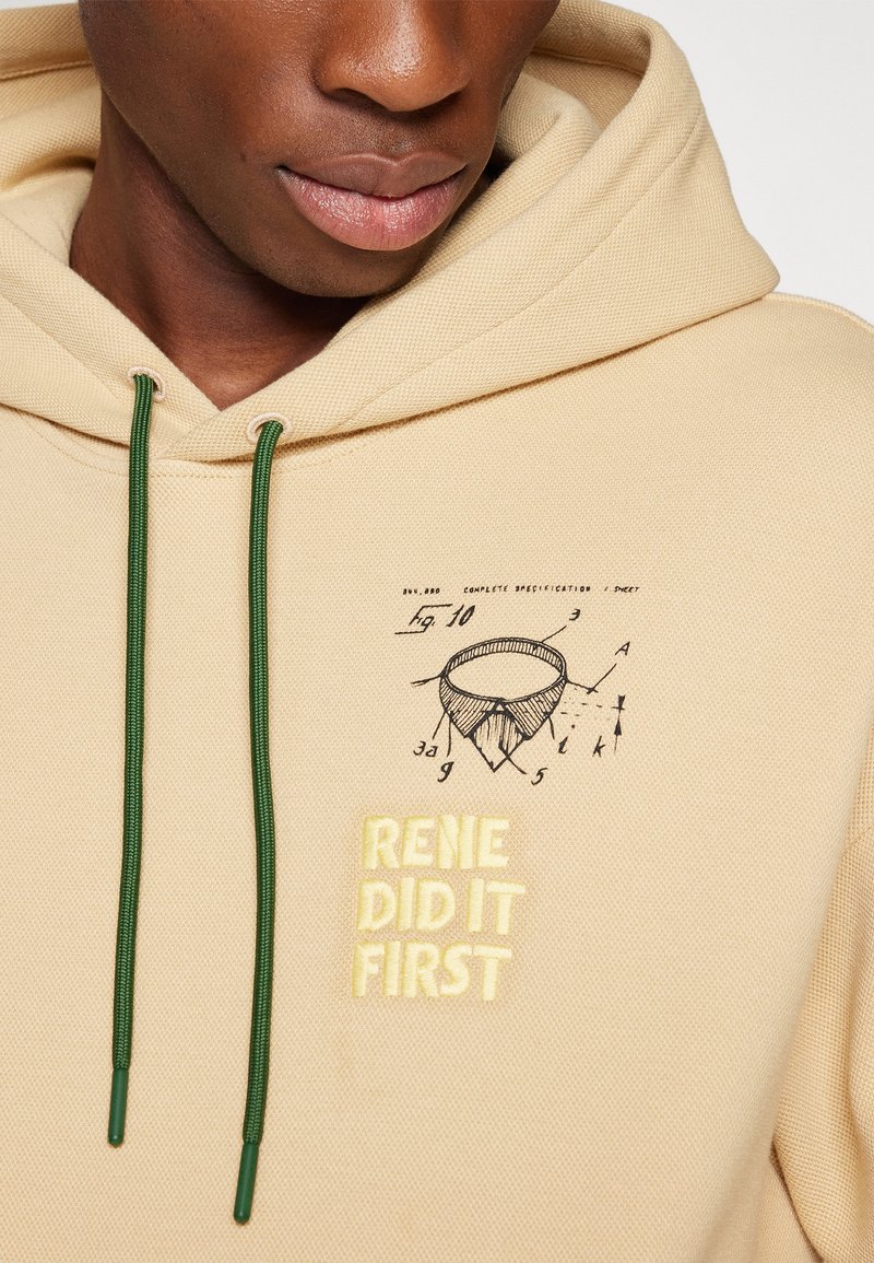 Beige hoodie met groene trekkoorden. Voorzien van een illustratie en de tekst "RENE DID IT FIRST" in het geel. Zacht, textured stof.