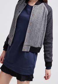 Woman wearing a navy blue dress with a black pleated hem and a grey textured buttoned bomber jacket with black cuffs and collar.