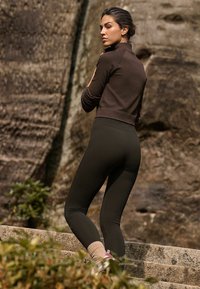 Brown ribbed long-sleeve top paired with high-waisted black leggings and light-colored socks. Outdoor setting with rocky backdrop.